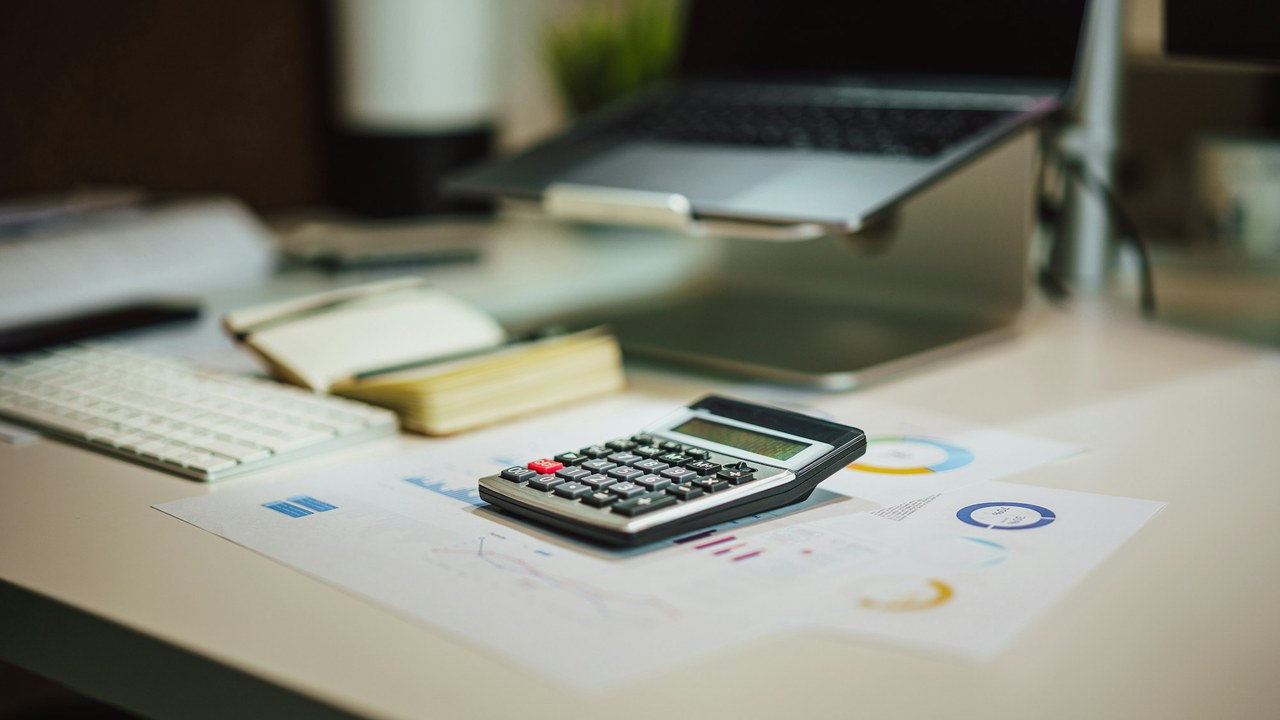 A calculator on printed charts at a desk, with a laptop and notebooks behind it.