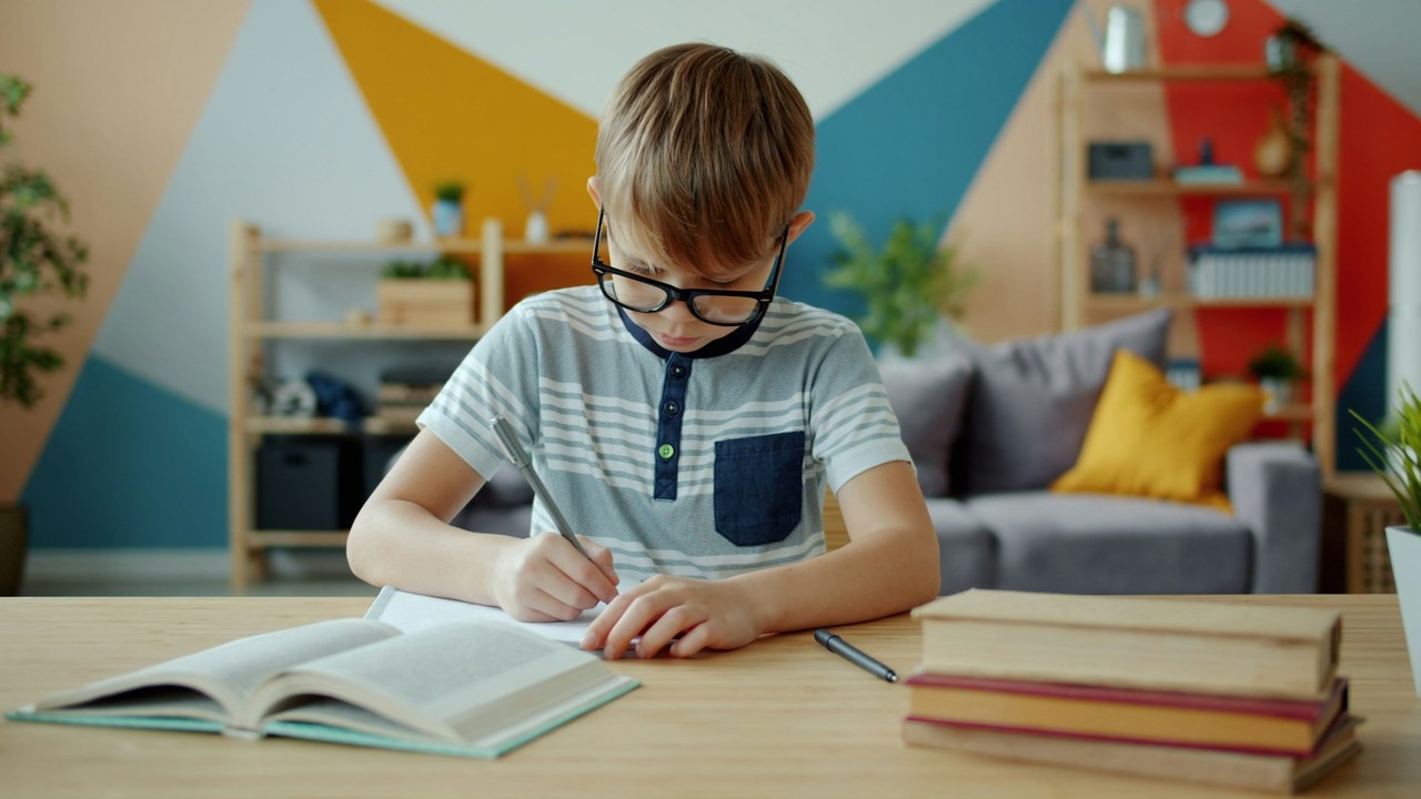 A boy in glasses writing in a notebook at his desk, a stack of books beside him.