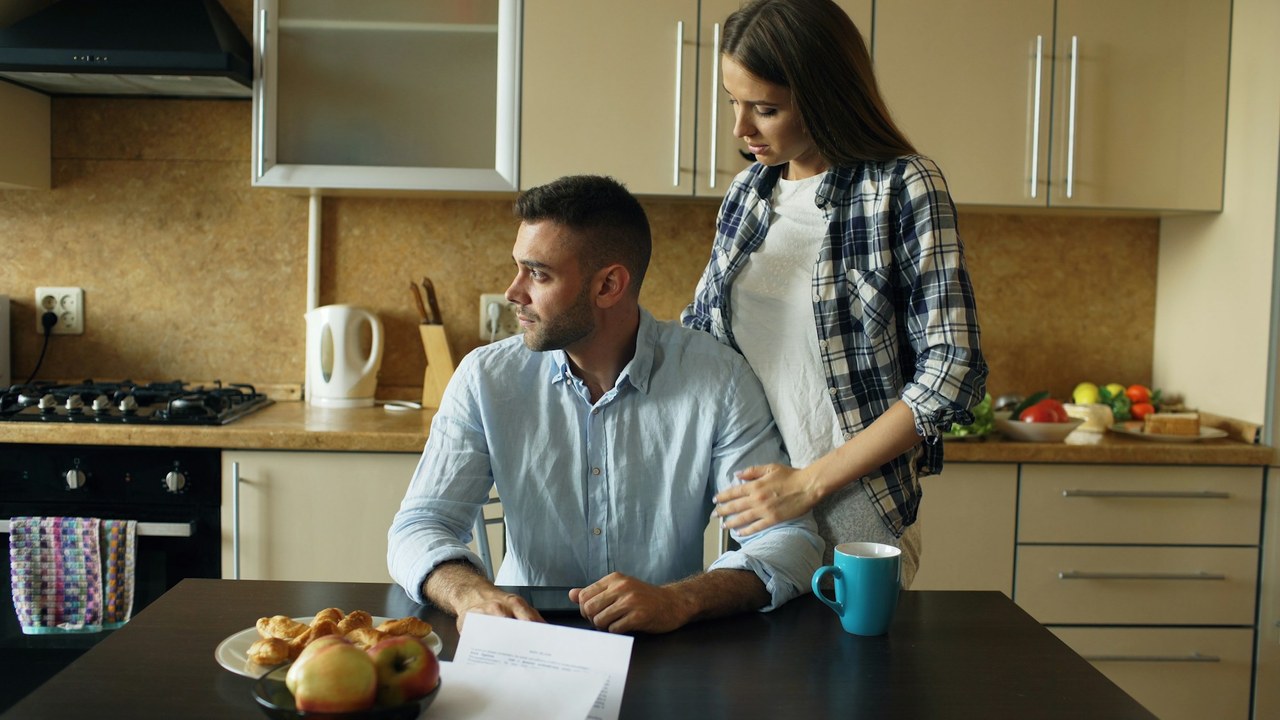 A couple in their kitchen, a sheet of paper on the table in front of them.