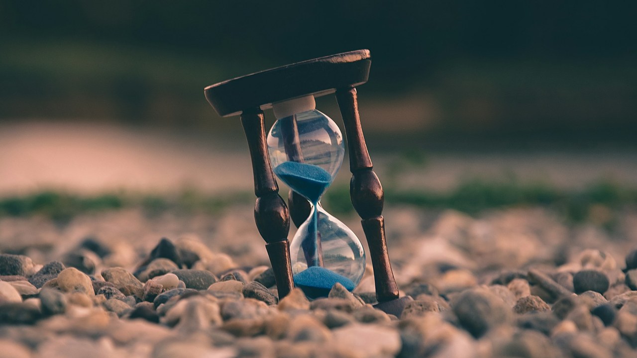 An hourglass with blue sand standing on gravel.