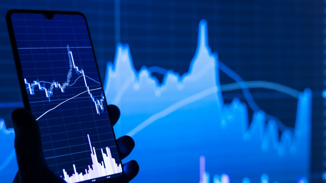 A phone showing a stock chart, held in front of a larger chart on a screen.