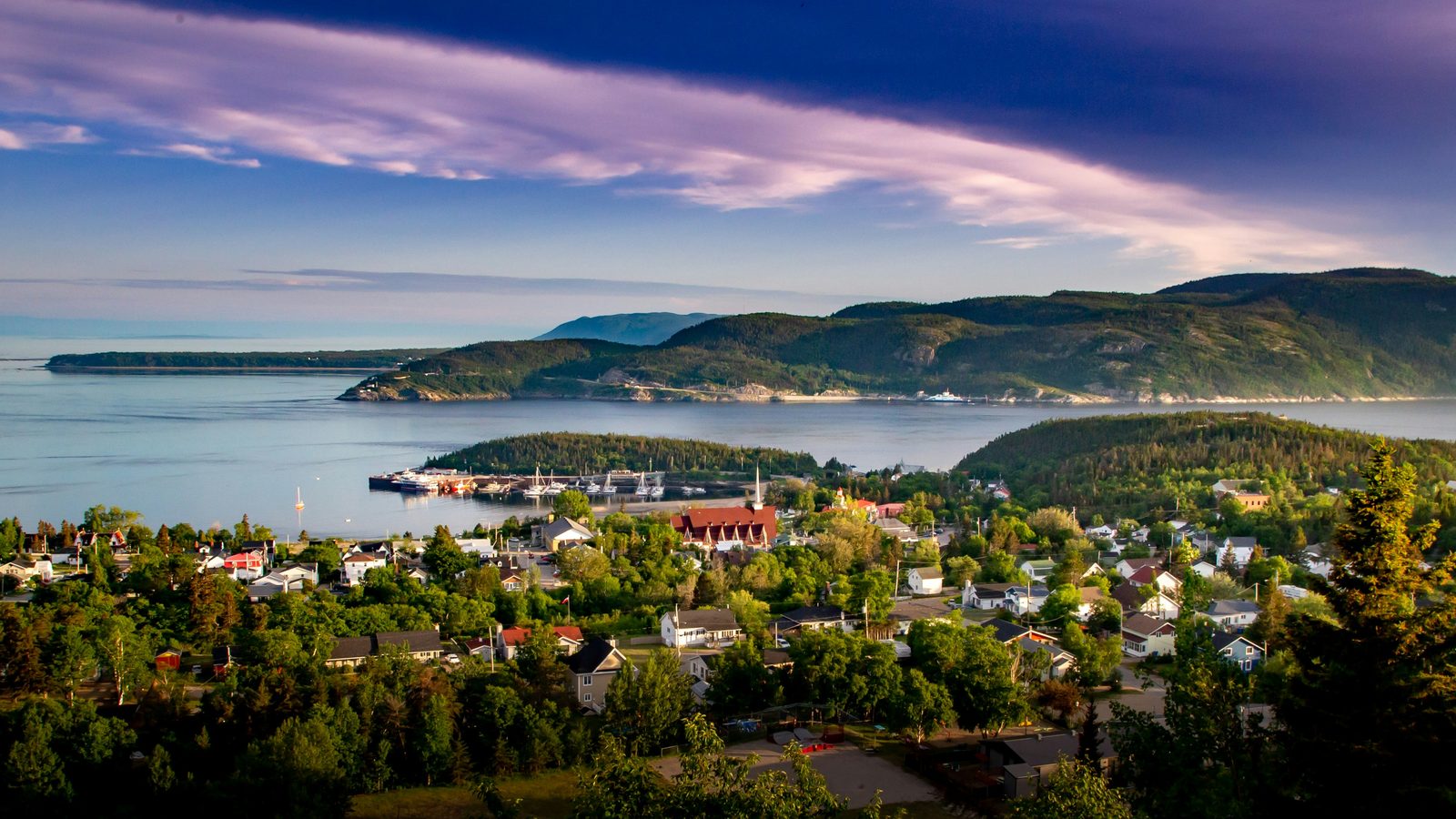 A small Québec coastal village nestled between wooded hills and the St. Lawrence River at golden hour.