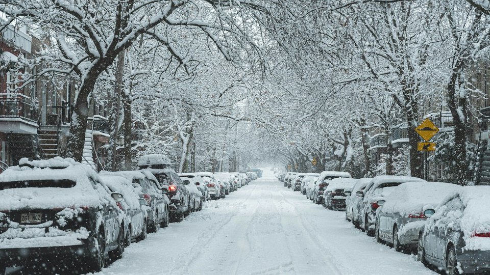 A residential street during a heavy snowfall, parked cars covered in snow along both sides.